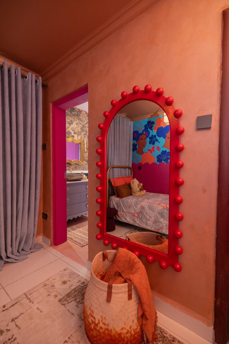 Arched mirror with red pom-pom trim in the hallway reflecting the bedroom, with an ensuite bathroom visible through an open pink-framed door